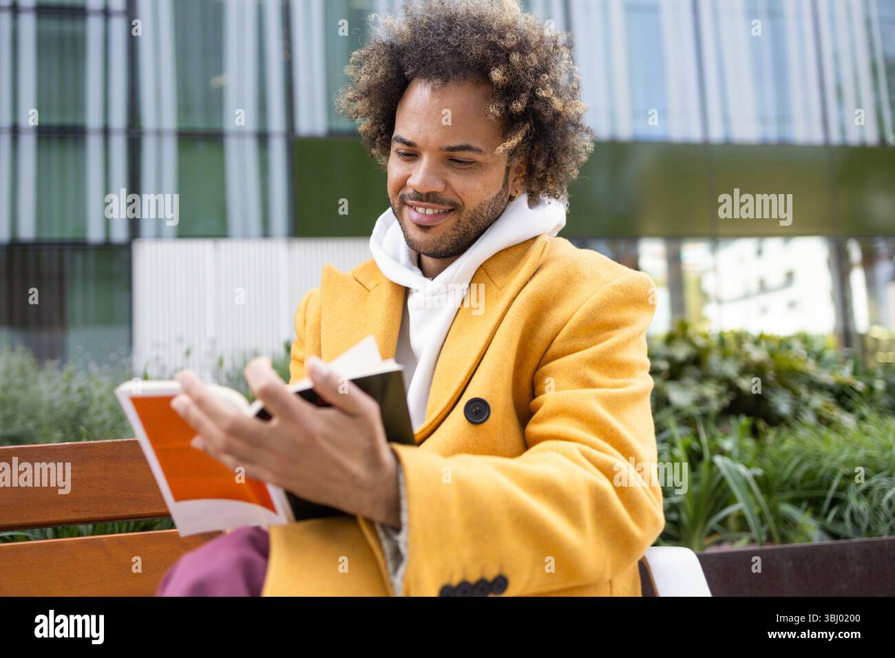 A cheerful Afro man with curly hair, wearing a bright yellow coat, enjoying a peaceful moment reading a book on a bench in an urban outdoor setting. Stock Photo