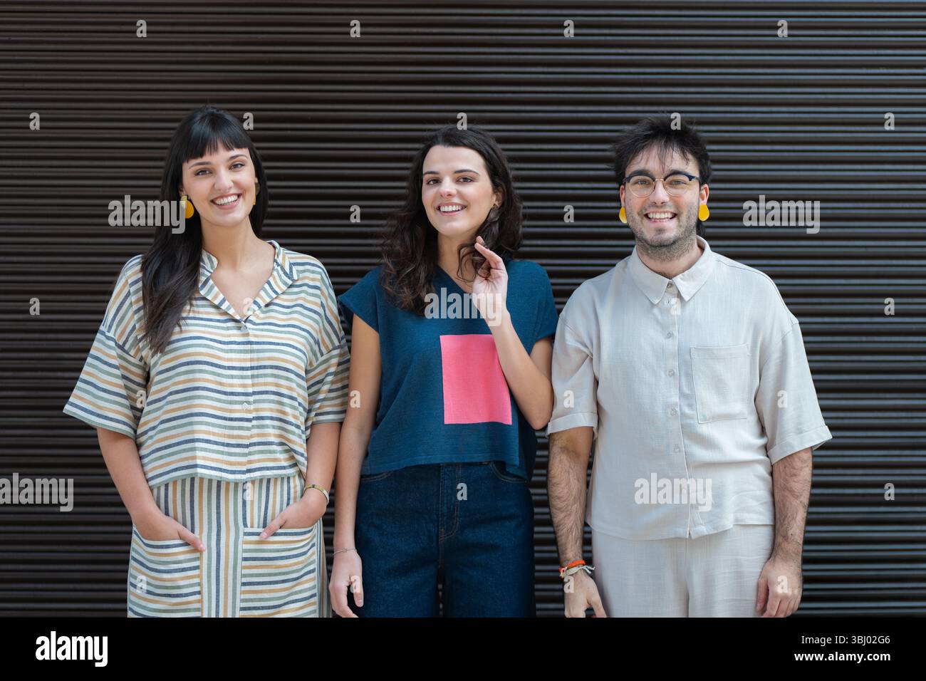 A diverse group of three friends stands together, smiling warmly at the camera. They each wear casual, trendy clothing while standing against a dark, Stock Photo