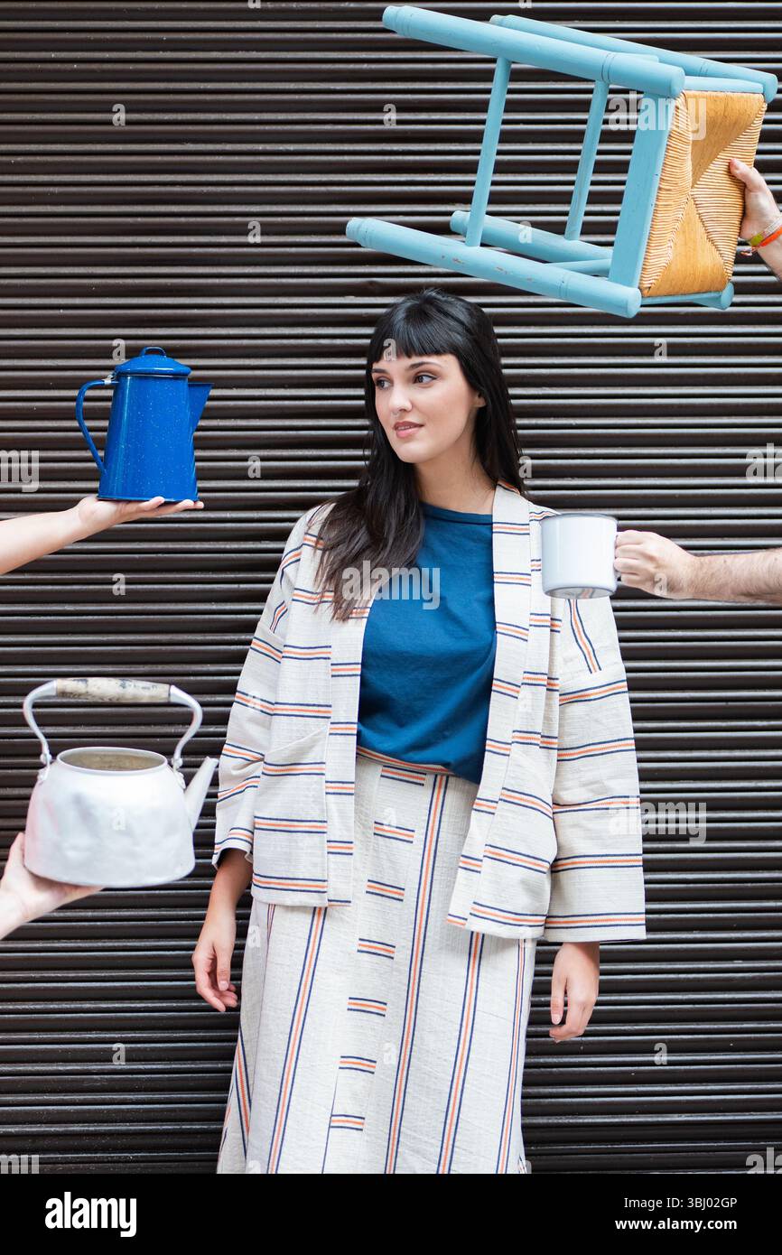 A stylish woman in a striped outfit poses in an urban setting, surrounded by a floating chair and kitchenware, blending fashion with surreal elements, Stock Photo