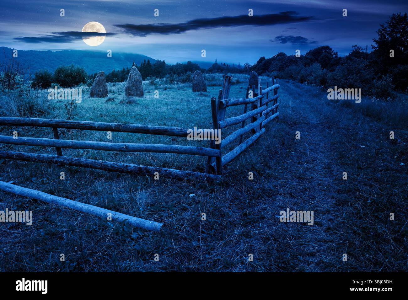 rural field behind the wooden fence at night. countryside landscape in mountains in full moon light. organic haystack on the meadow Stock Photo