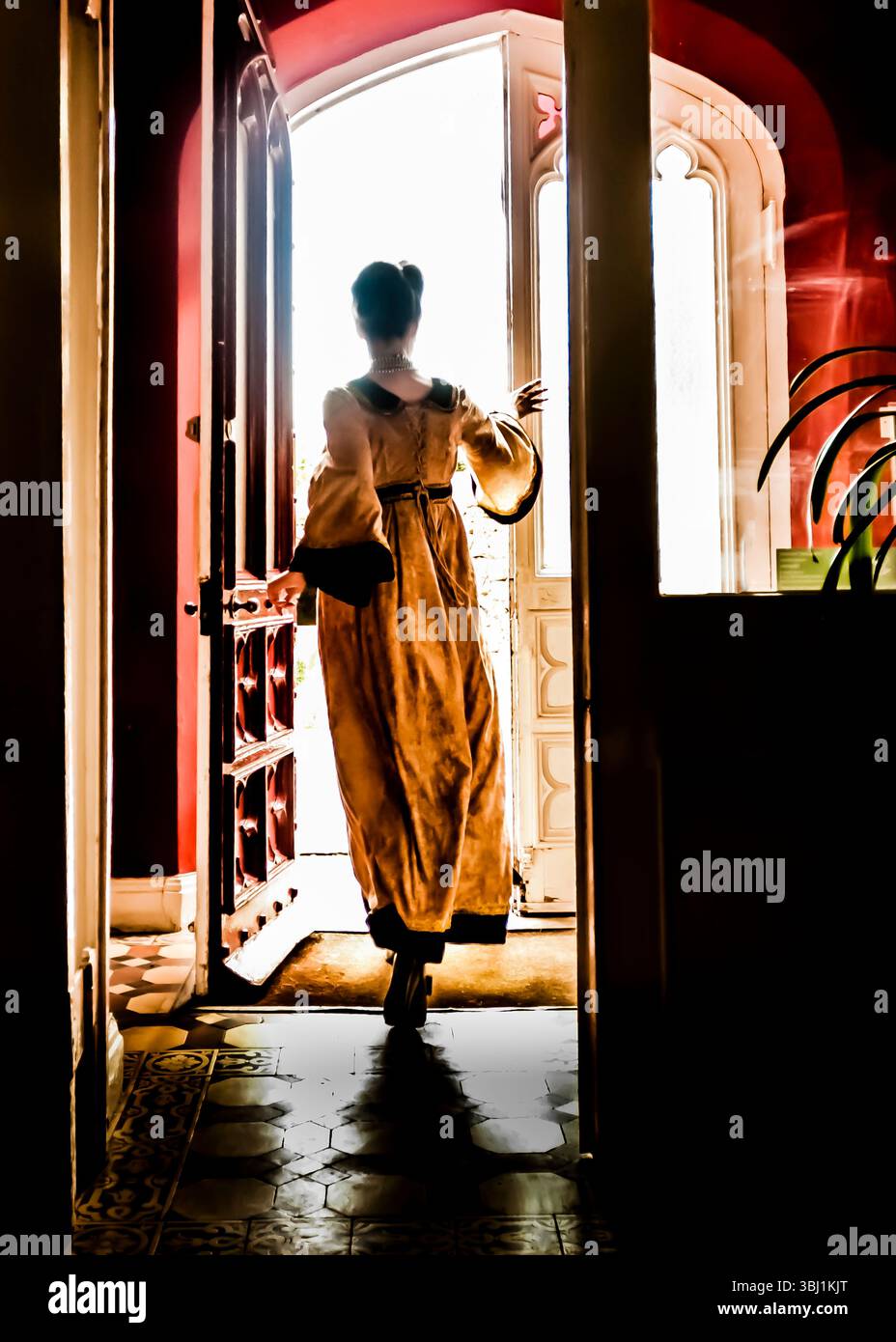A woman in a long dress is silhouetted in a doorway, with the bright light of the outside world illuminating her from behind. Stock Photo