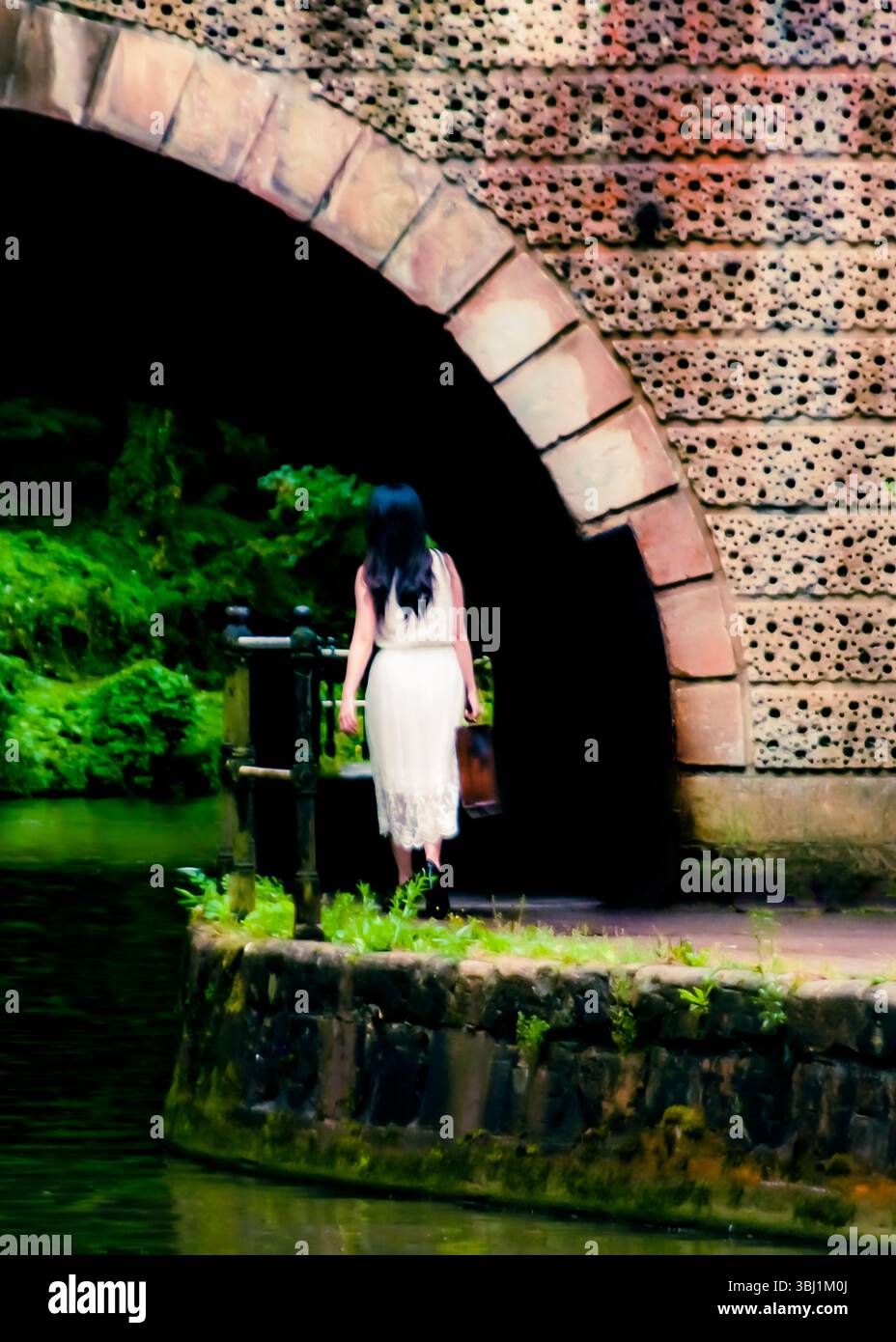 A woman in a white dress walks under an archway, carrying a brown bag. Stock Photo
