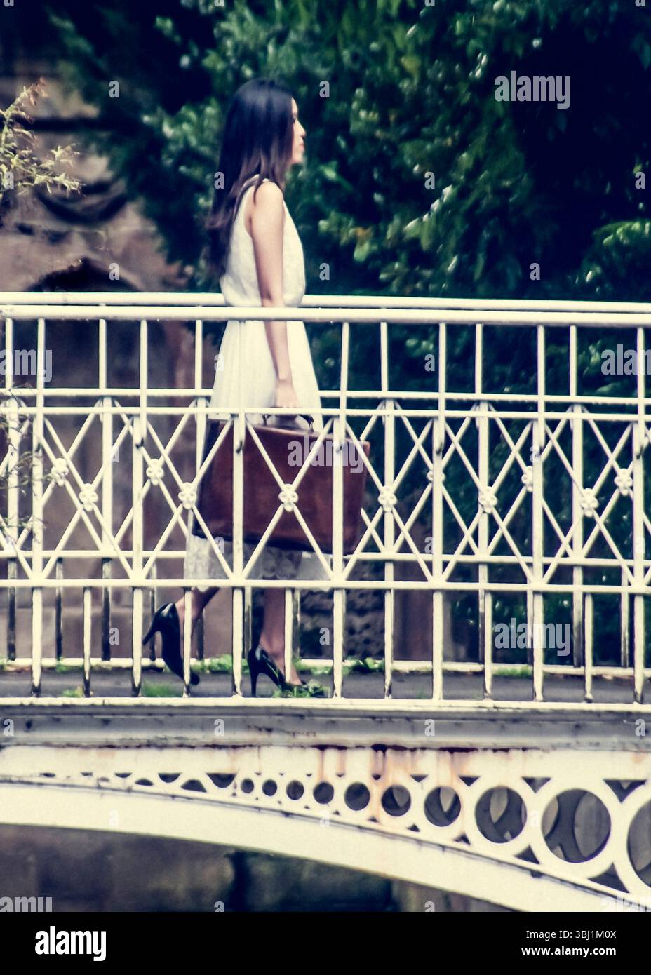 A woman in a white dress and heels walks across a bridge, carrying a suitcase. Stock Photo