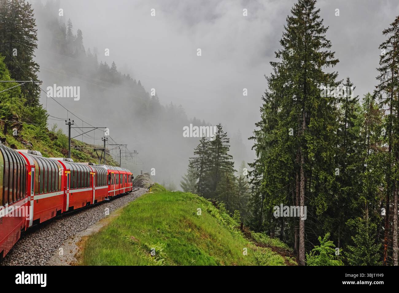 A red train winds along a narrow track amidst a misty mountain landscape in Switzerland Stock Photo