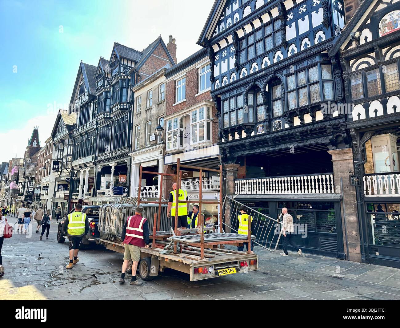Workers in high-vis vests load metal fencing onto open-back truck after Chester Half Marathon on Eastgate Street, sunny day - Smartphone Captured Stock Image