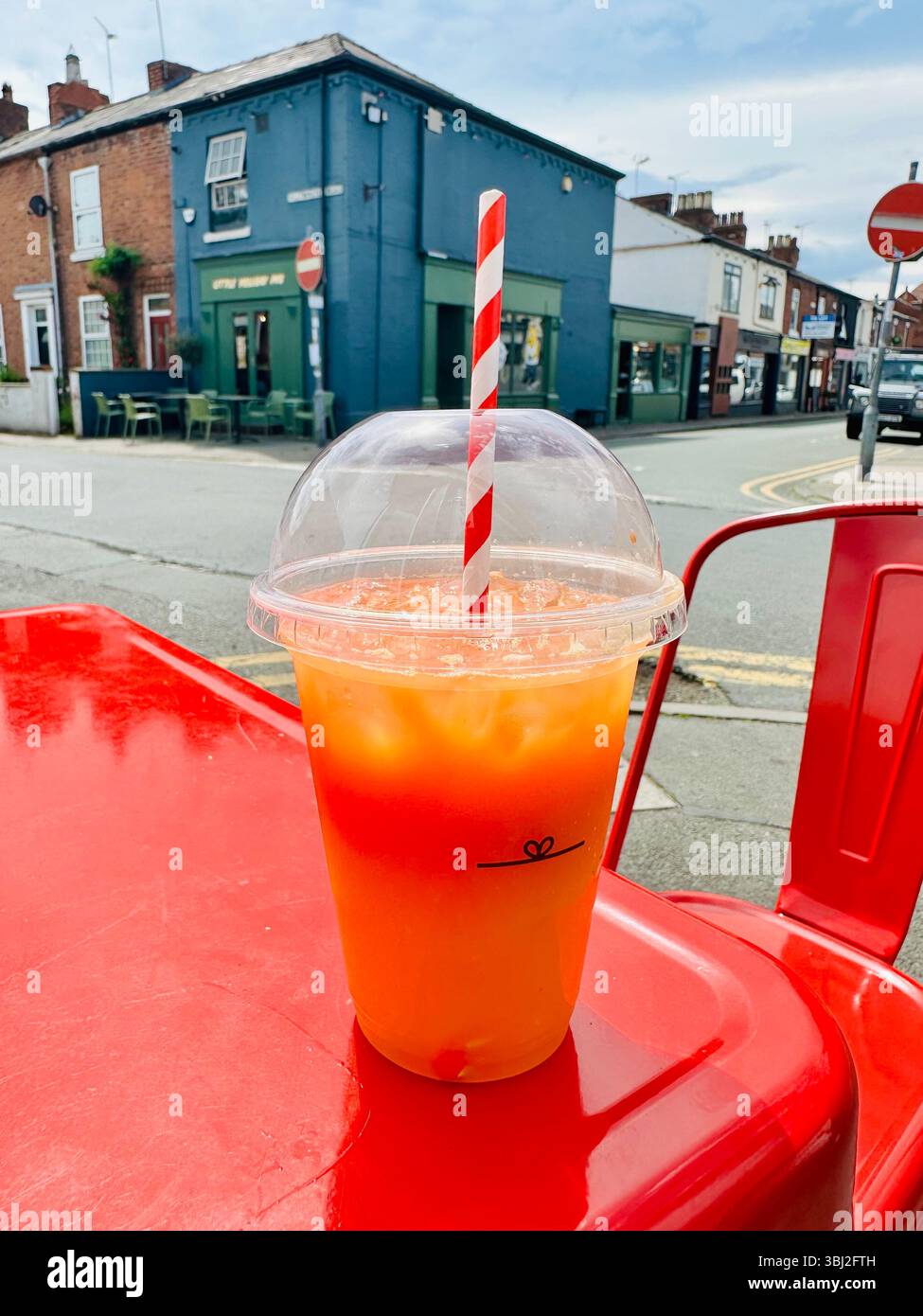 Orange carrot and ginger juice in takeaway cup with striped straw on red metal table, street corner and local café visible in background - Smartphone Captured Stock Image