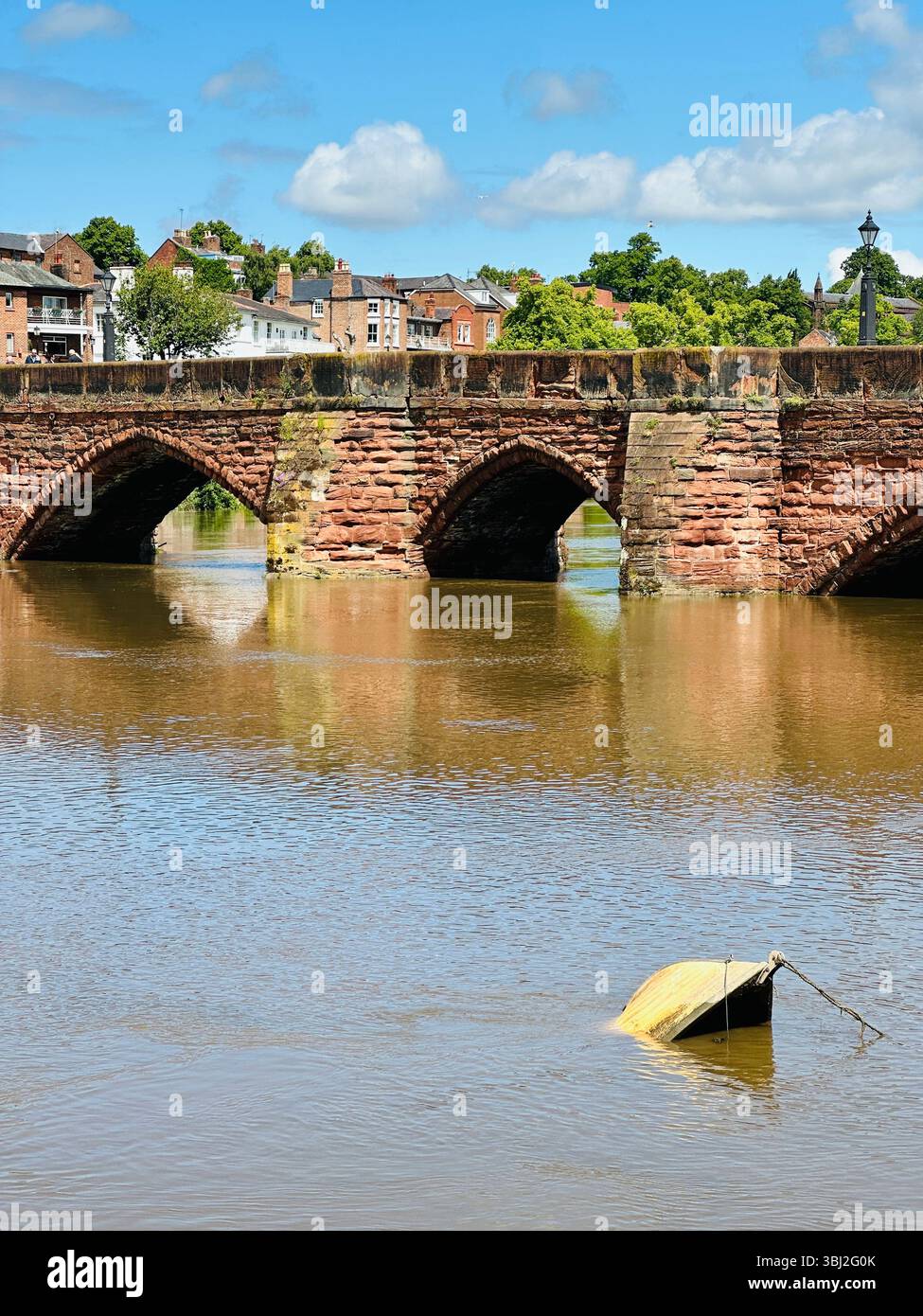 View of River Dee in Chester with red brick bridge to Handbridge, small green capsized boat, and blue sky on a sunny day - Smartphone Captured Stock Image
