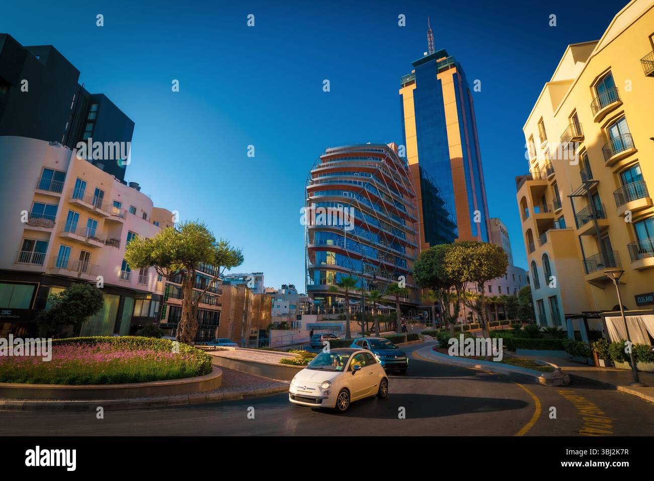 Scenic view of a white Fiat 500 car driving around a roundabout in front of the Portomaso Business Tower in Saint Julian's (San Ġiljan), Malta Stock Photo