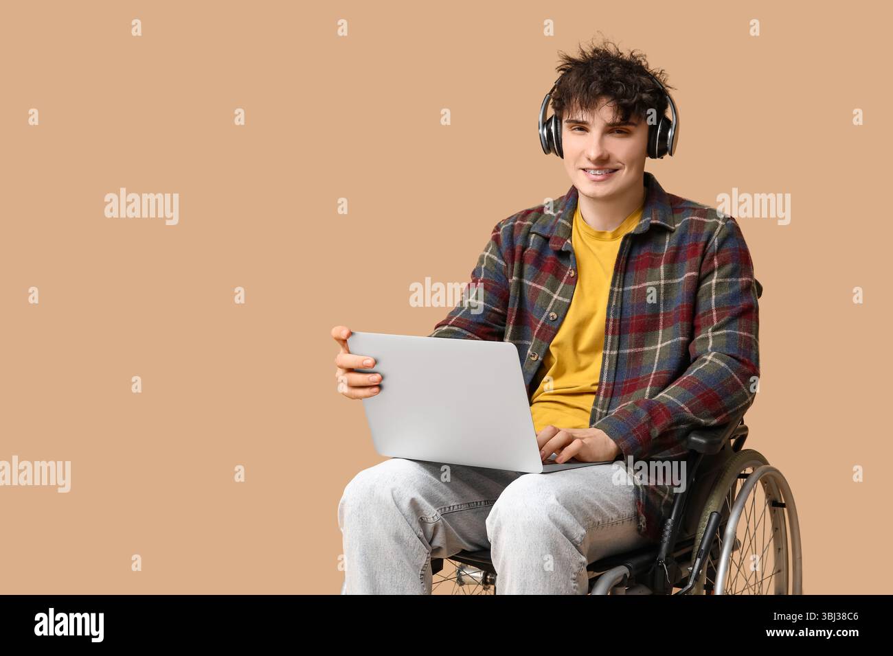 Young male programmer in headphones on wheelchair holding laptop on beige background Stock Photo