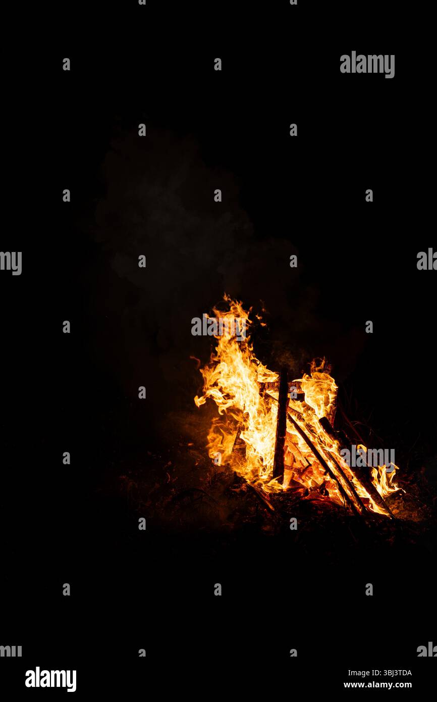 Wooden bonfire flames rise in the dark night, creating a warm glow and dramatic contrast against the black background. A traditional scene from the Sa Stock Photo