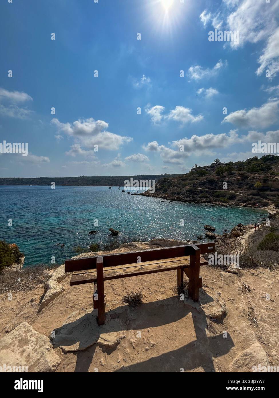 A wooden bench on a rocky cliff overlooks the clear turquoise sea under a bright sunny sky with scattered clouds, offering a peaceful coastal view. - Smartphone Captured Stock Image