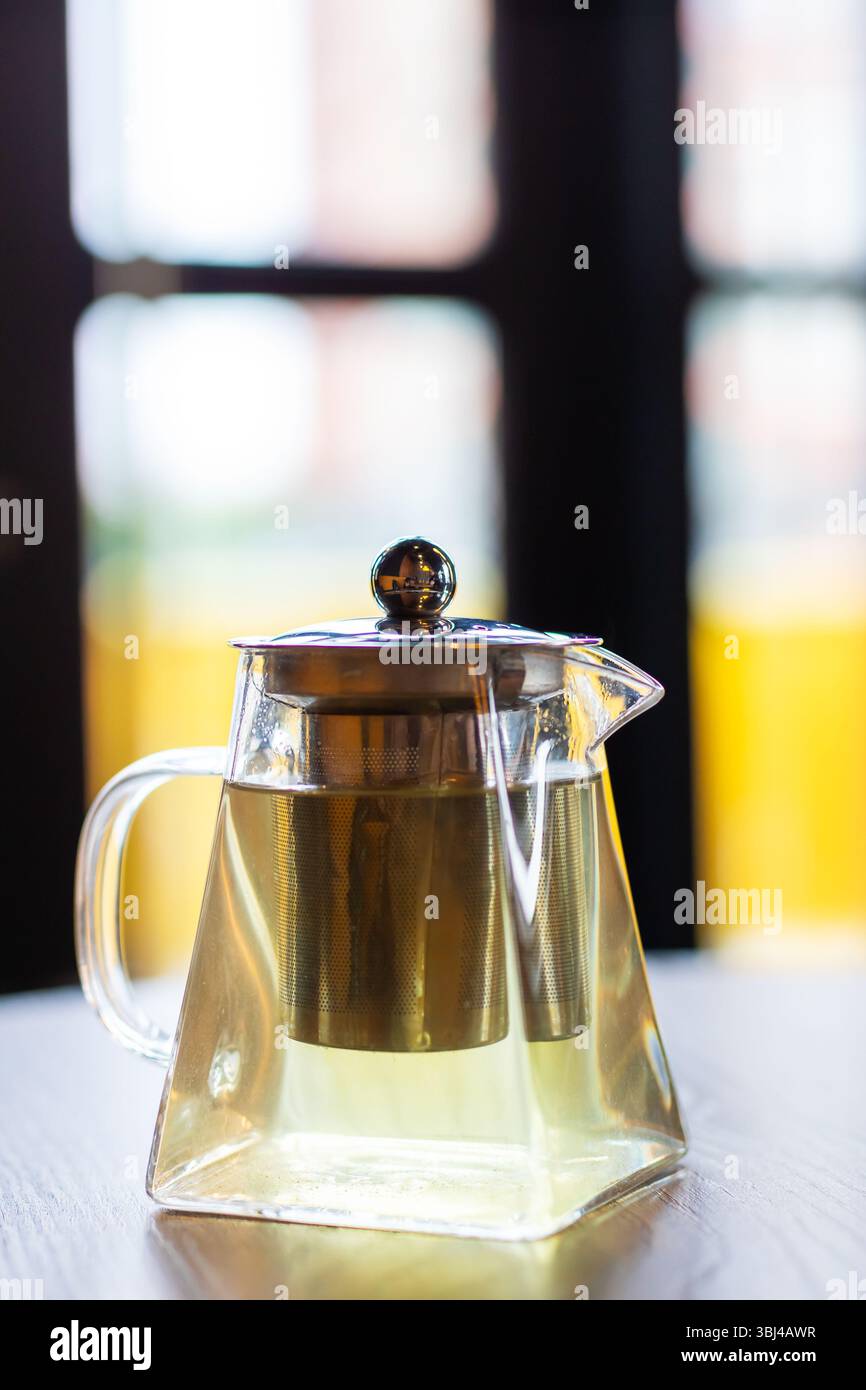 On a table, there is a beautiful glass teapot that features a brass filter, elegantly displayed alongside various serveware items Stock Photo