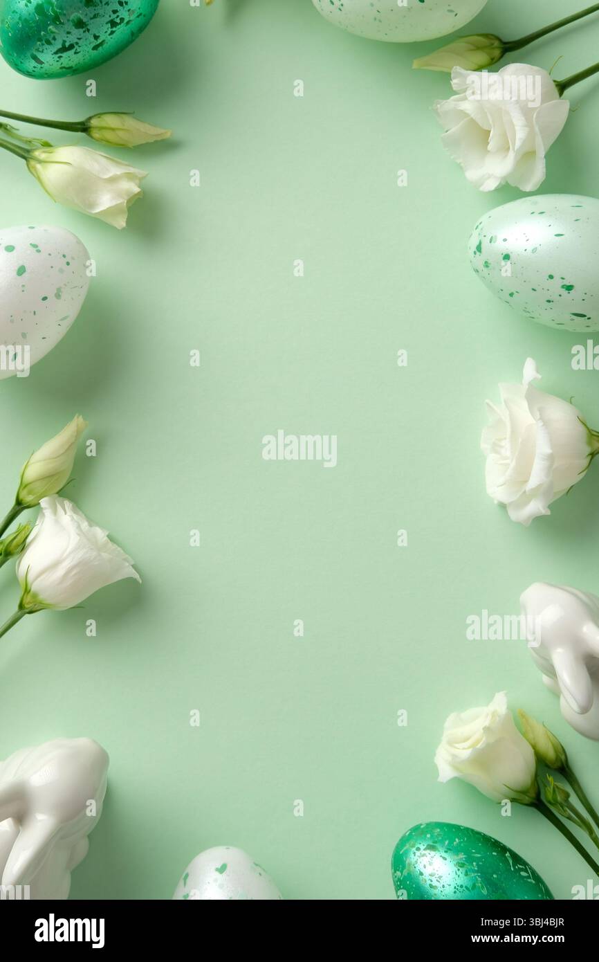 Easter flat lay composition with speckled eggs, white spring flowers, and pastel ceramic bunnies on a soft green background Stock Photo