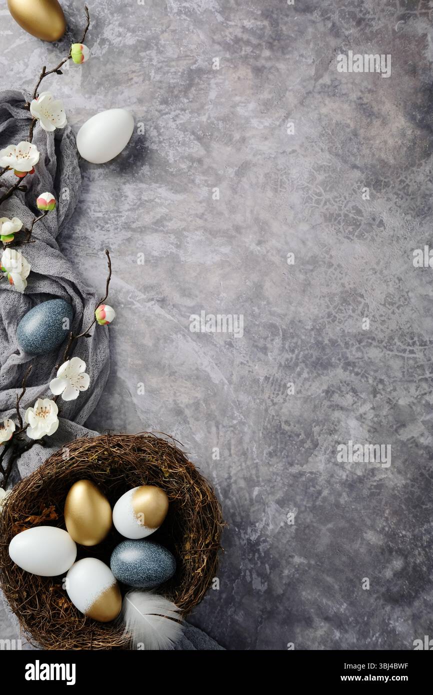 Easter flat lay with a nest of gold, white, and blue decorated eggs, blooming branches, and a grey cloth on a textured grey background Stock Photo