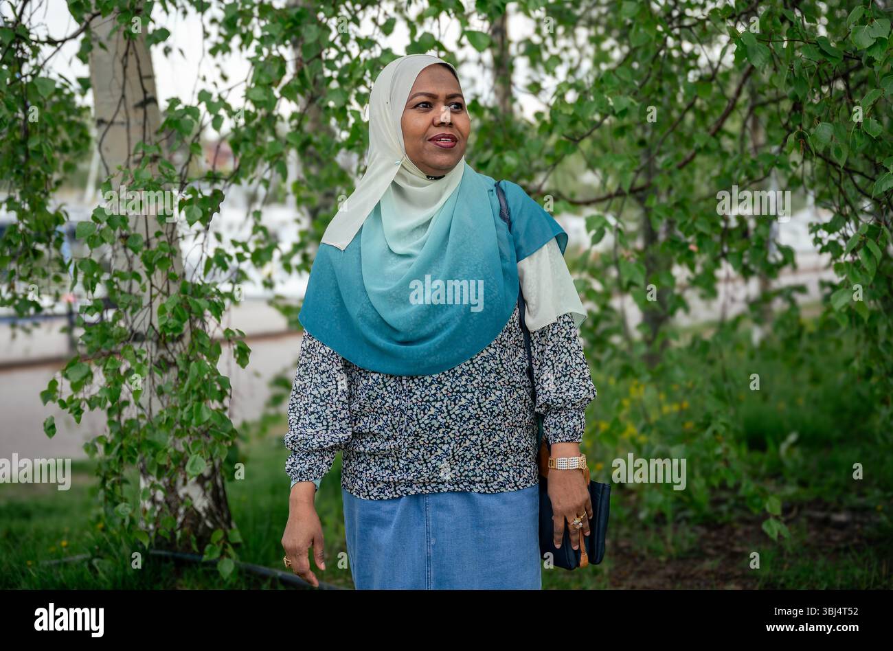 woman in a teal hijab and blouse standing outdoors under leafy trees Stock Photo