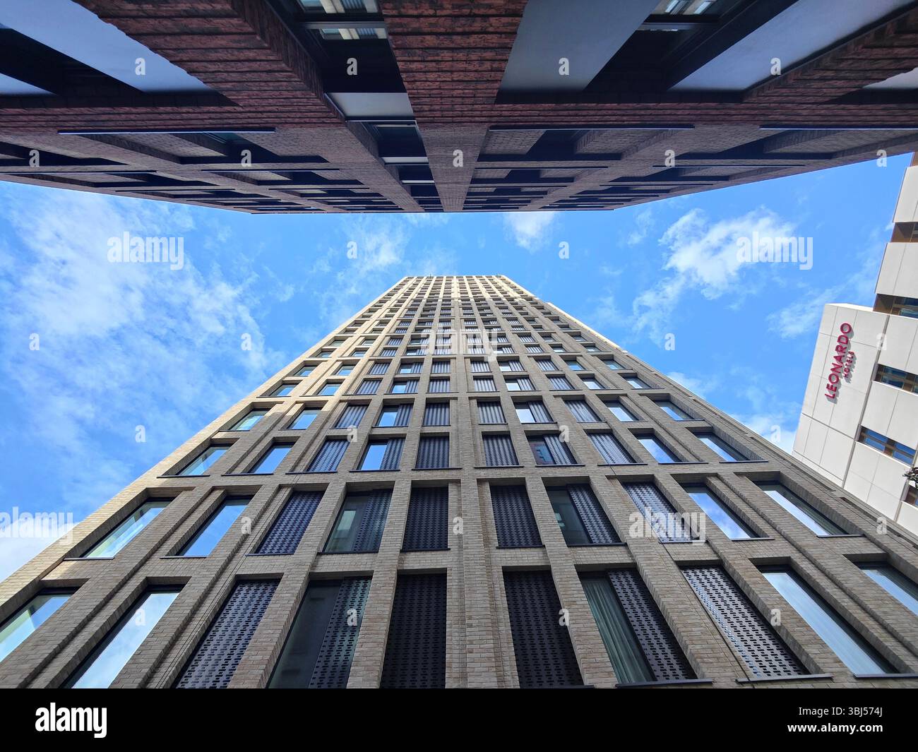 Upward view of a modern skyscraper with geometric patterns against a vibrant blue sky, juxtaposed with neighbouring buildings and scattered clouds. - Smartphone Captured Stock Image
