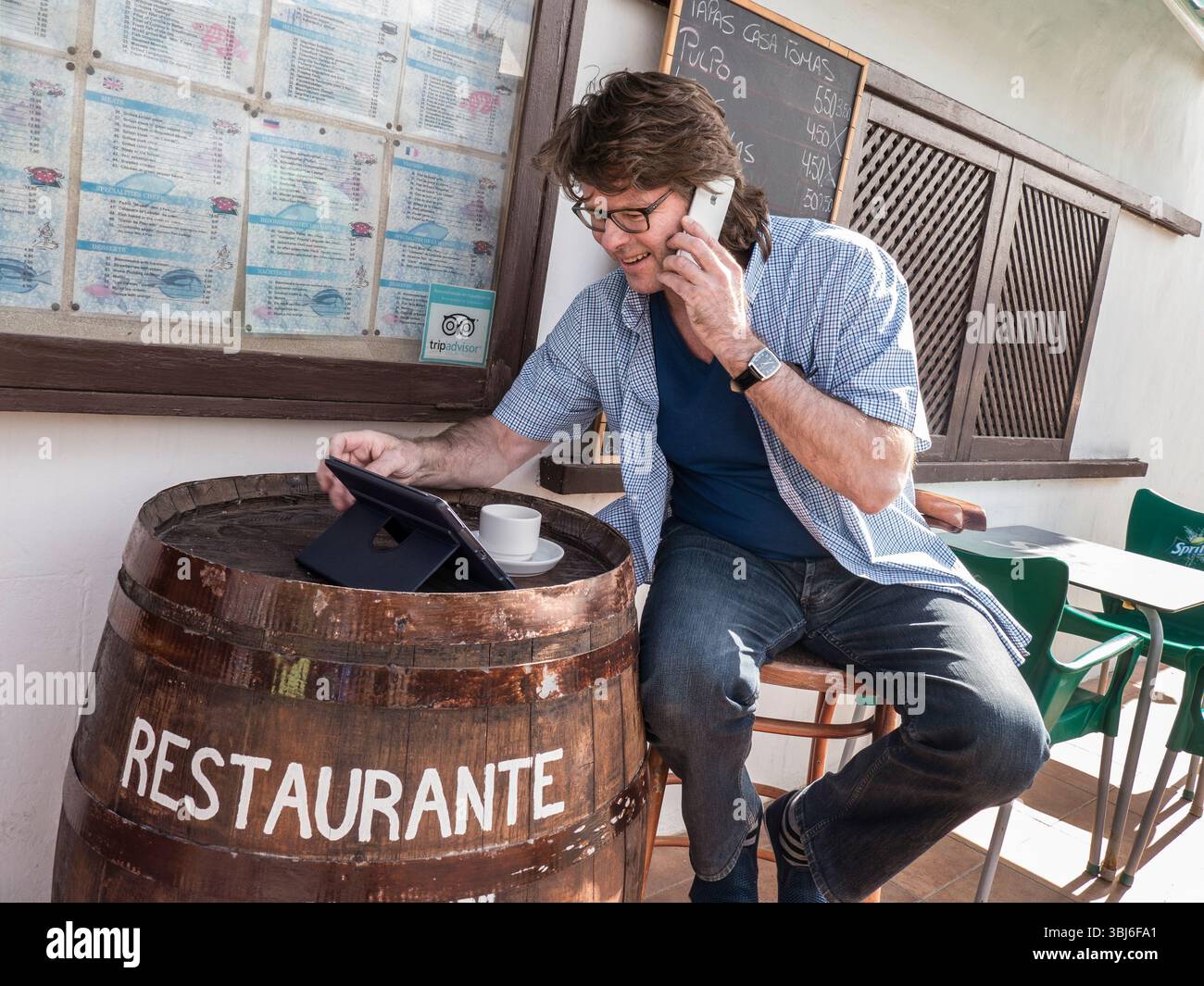 Mature businessman using his iPhone smartphone & iPad Air tablet computer abroad alfresco Spanish Tapas restaurant bar, international communications. Stock Photo