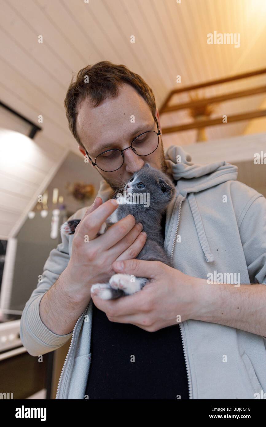 A heartwarming scene of a man lovingly holding a gray kitten, sharing a tender moment in a warm and inviting home filled with cozy decor and natural l Stock Photo