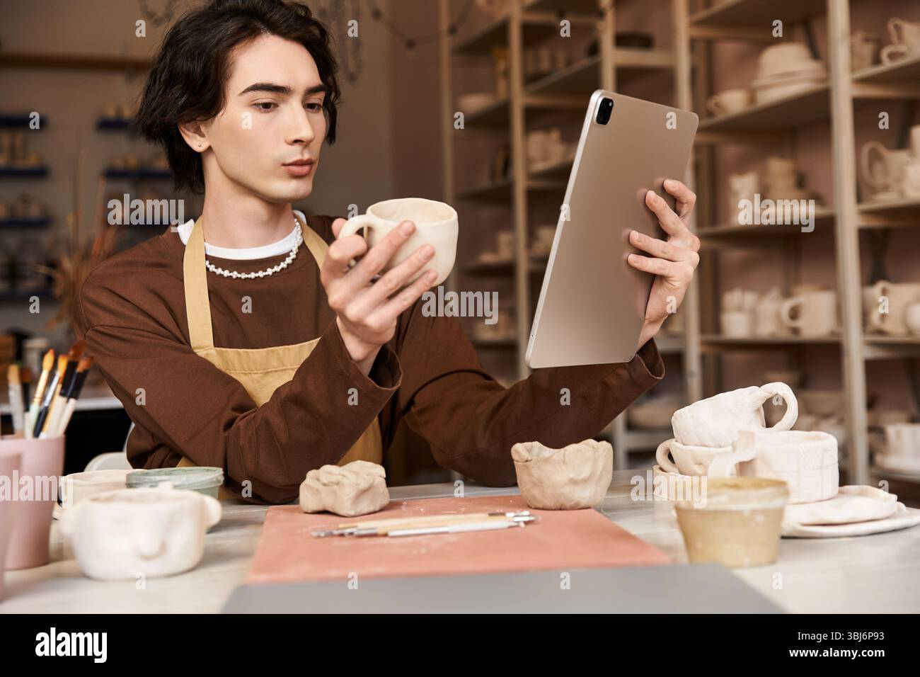 A man in a pottery studio examines a clay mug while using a tablet for creative inspiration. Stock Photo