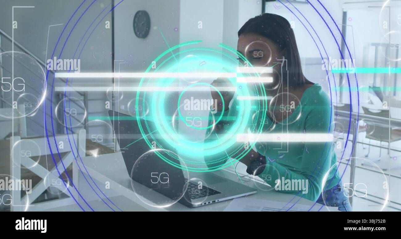 Holding mug, mid adult woman typing on silver laptop at open-plan office, with 5G interface Stock Photo