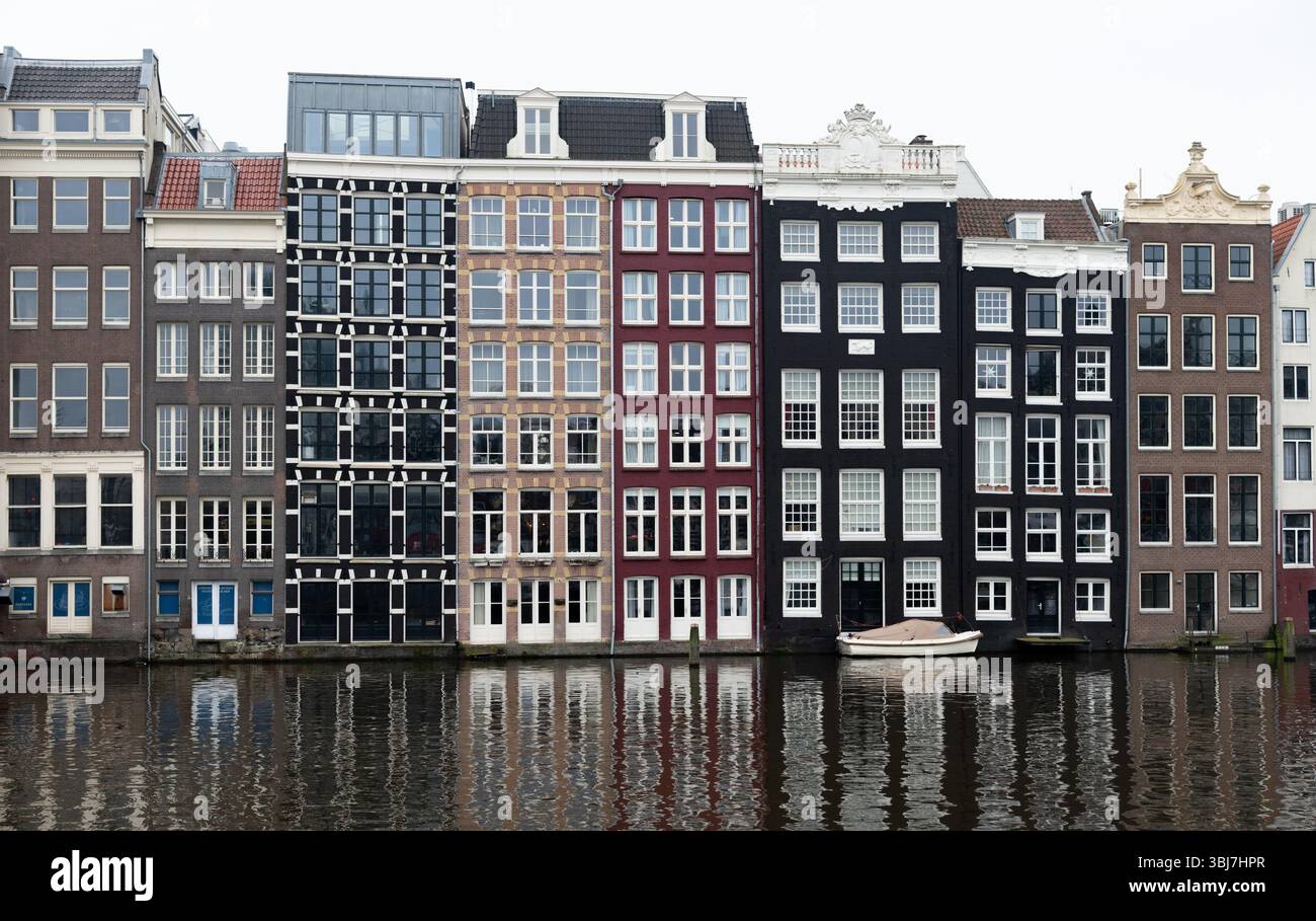 Row of colorful narrow buildings reflecting on a canal in amsterdam, netherlands, with a small boat moored in front, on a cloudy day Stock Photo