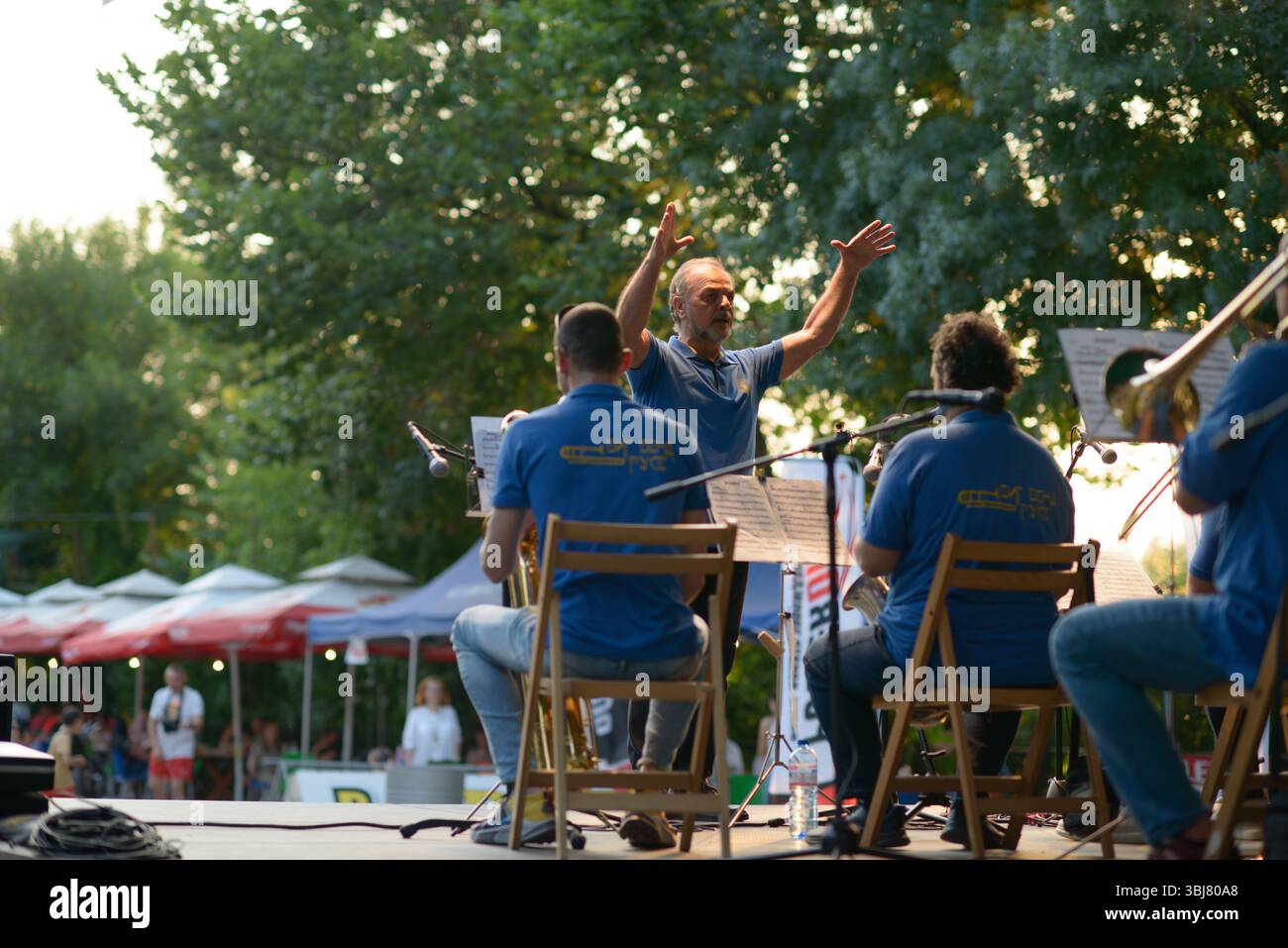 A view from behind the stage shows the conductor of Big Band Ruse leading a live performance during the Moto Rock Fest 2025 in Ruse, Bulgaria, on 13 June. The musicians, dressed in matching blue shirts, perform under a canopy of trees and stage lights, with sheet music and microphones visible. The conductor raises his arms in expressive motion as the crowd gathers in the distance under rows of festival umbrellas. The open-air concert blends classical and jazz traditions with the broader biker culture of the event, drawing families, tourists, and music lovers from across the region. Stock Photo