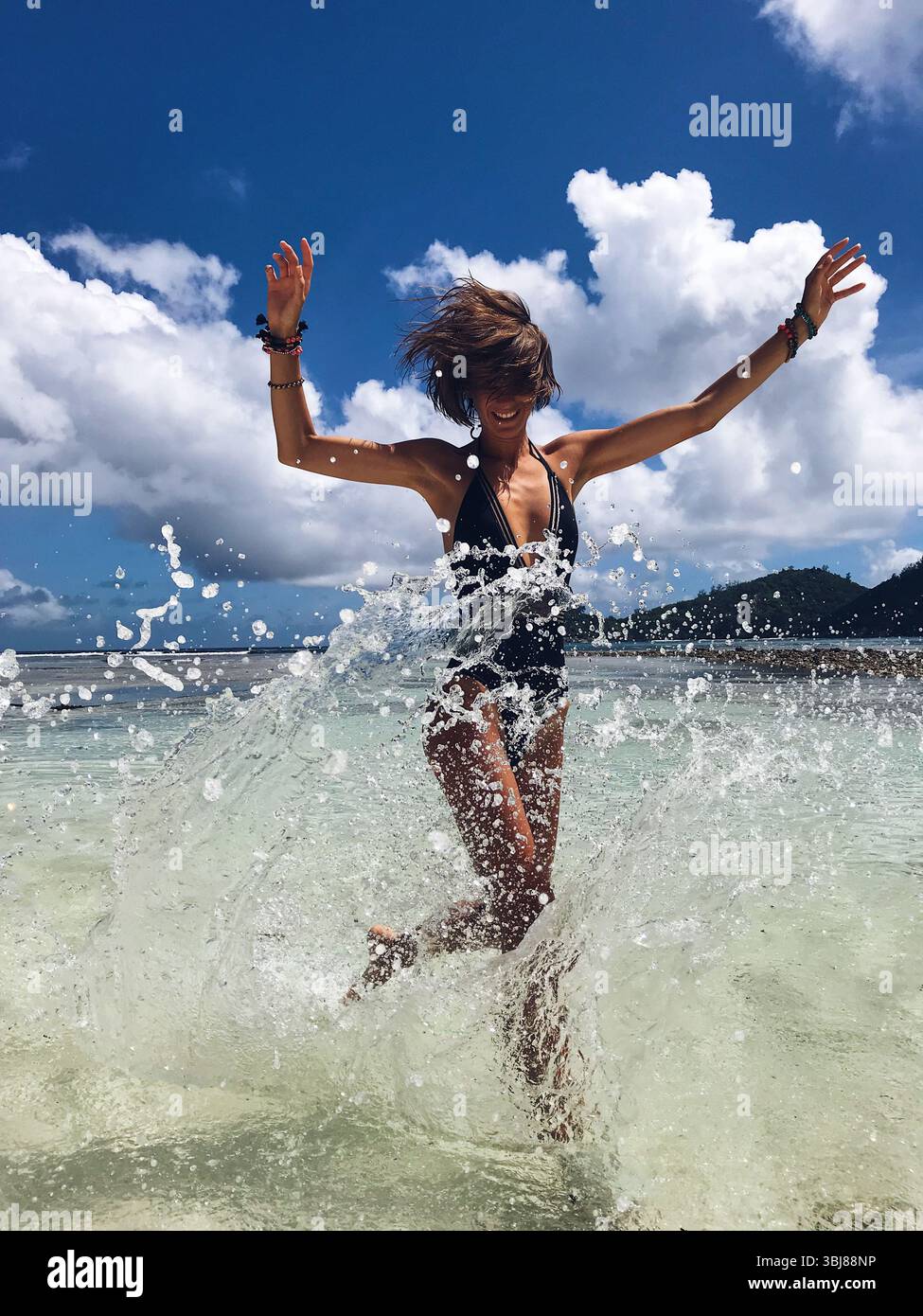 Beautiful woman running towards camera, while splashing water around with raised arms above head on the dream beach in Seychelles. Fashion model Stock Photo