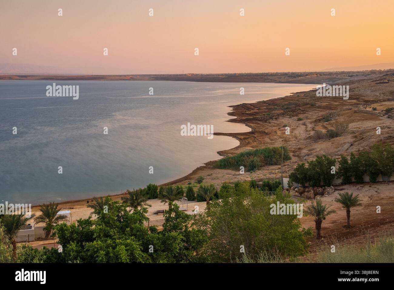 Sunset view over the Dead Sea coastline in Jordan, with palm trees, calm waters, and a serene desert landscape under golden light. Stock Photo