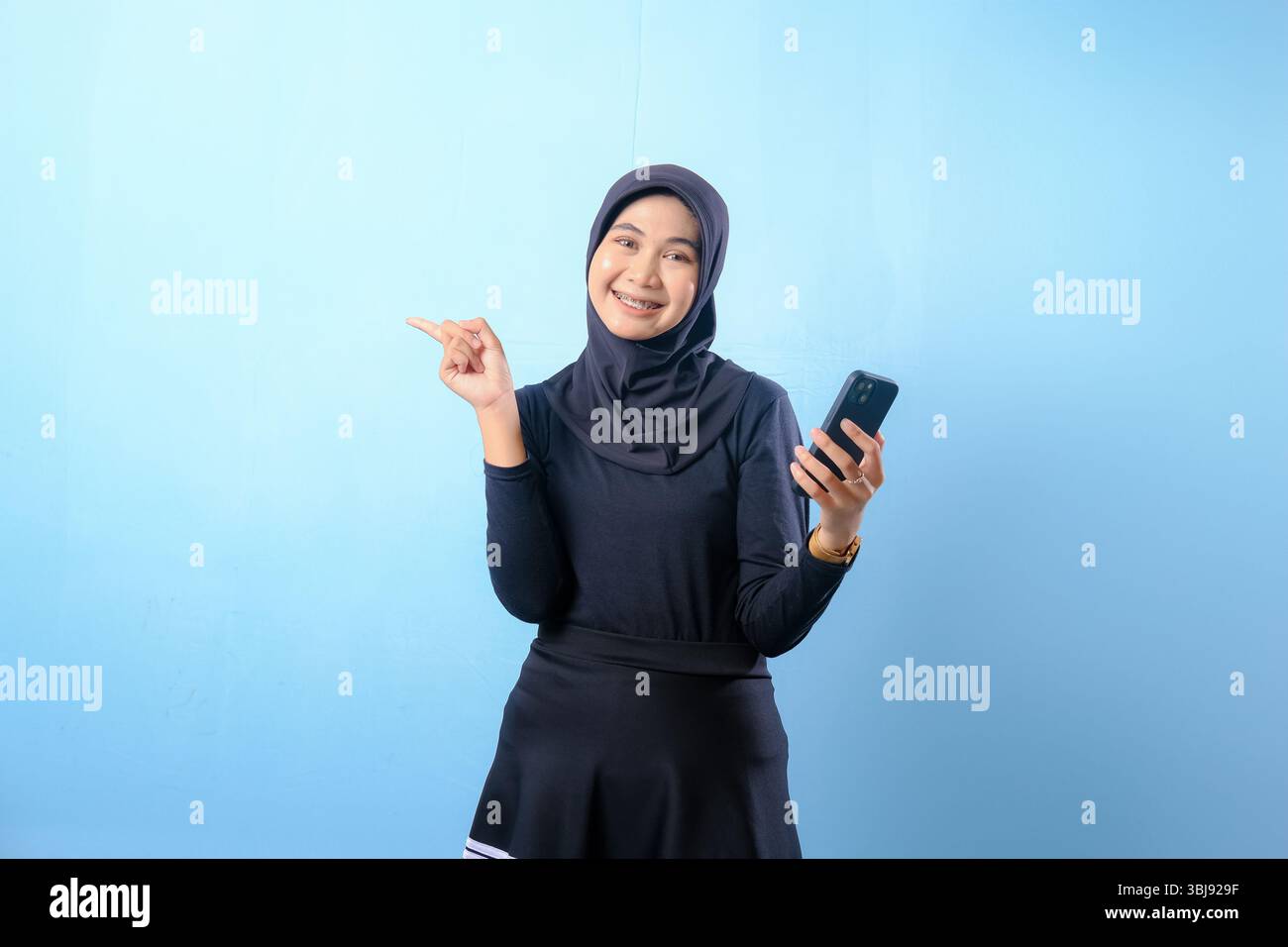 A woman wearing a black hijab is holding a cell phone and pointing to the camera. She is smiling and she is happy Stock Photo
