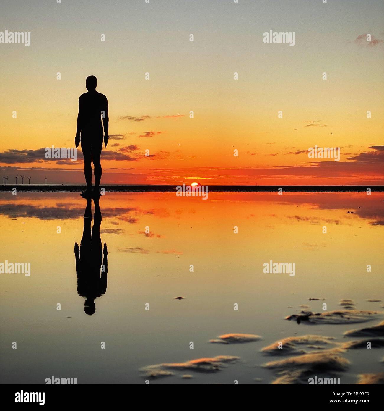 One of Anthony Gormley's Another Place Iron Man statues and vivid red sunset sky at Crosby beach reflected in a calm clear tide pool - Smartphone Captured Stock Image