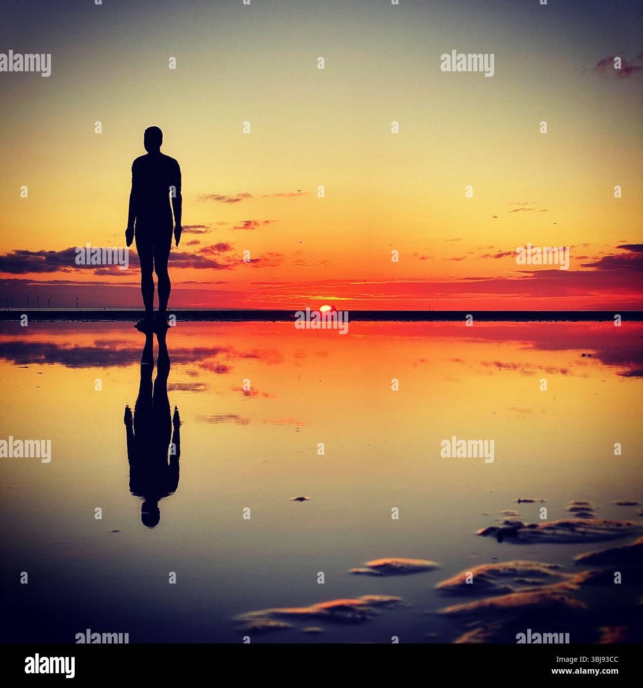 One of Anthony Gormley's Another Place Iron Man statues and vivid red sunset sky at Crosby beach reflected in a calm clear tide pool - Smartphone Captured Stock Image
