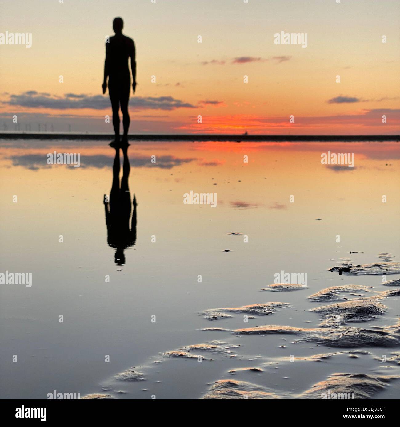 One of Anthony Gormley's Another Place Iron Man statues and vivid red sunset sky at Crosby beach reflected in a calm clear tide pool - Smartphone Captured Stock Image
