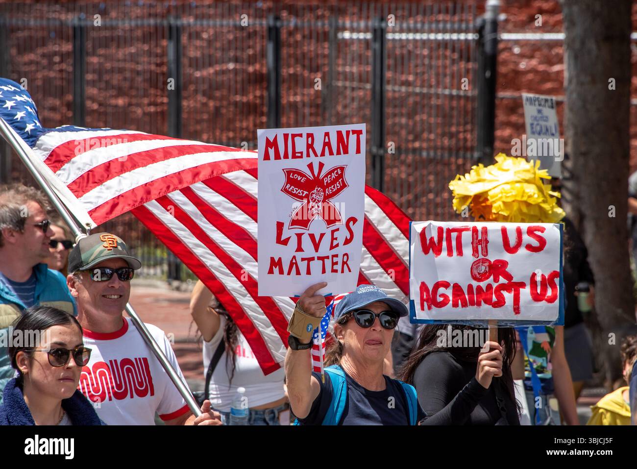San Francisco, USA. 14th June 2025. Among the protesters at the No Kings Day protest march, a man carries an American flag while women besidie him hold red and white signs reading, 'Migrant lives matter,' and 'With us or against us.' Credit: Shelly Rivoli/Alamy Live News - Stock Image