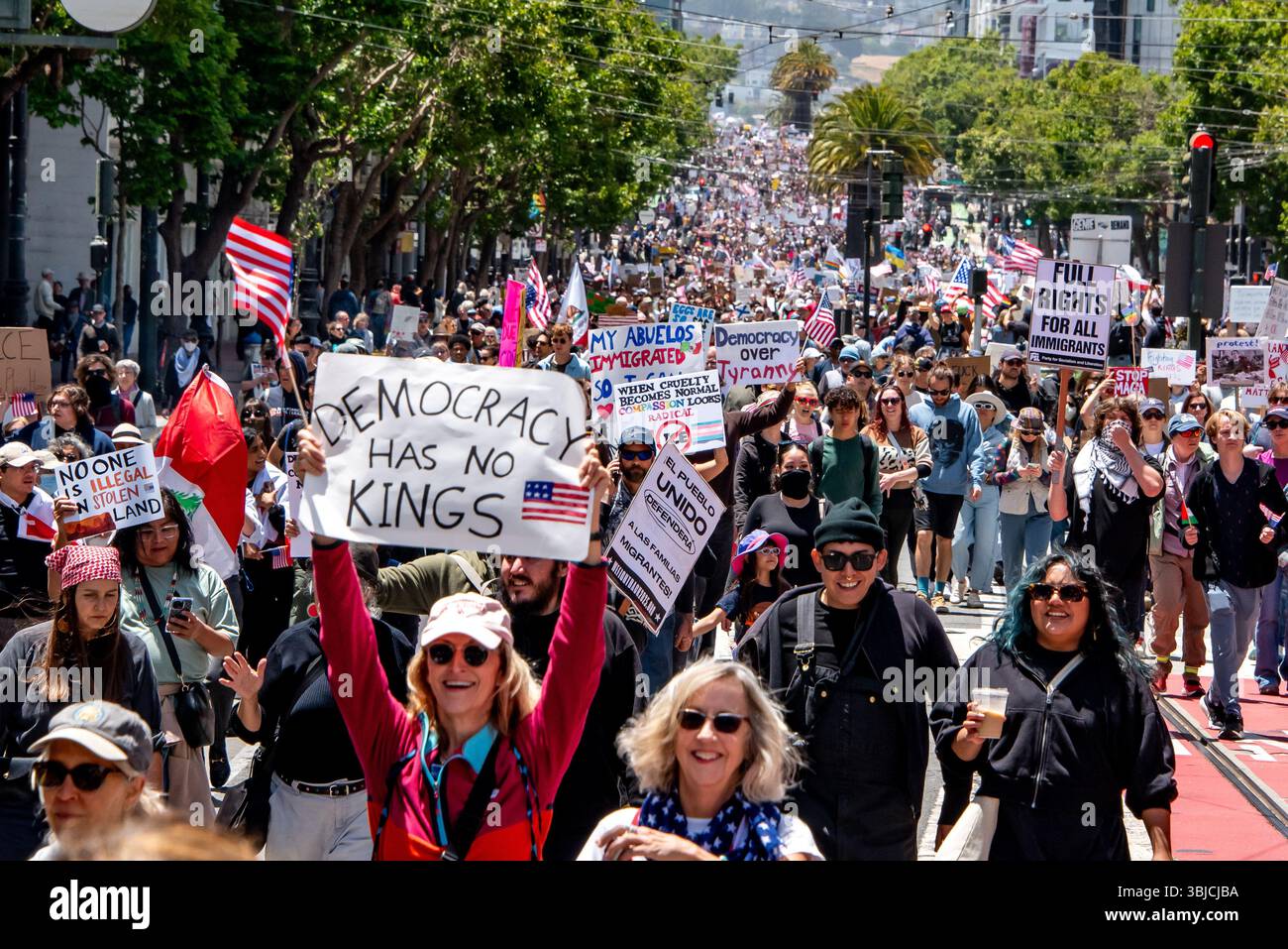 San Francisco, USA. 14th June 2025. As a crowd of thousands of protesters march down Market Street in the No Kings Day protest, many people hod signs high, including one in front reading, 'Democracy has no kings' held by a smiling woman with an American flag. Other signs seen address immigrant rights, democracy over tyranny, with many American flags and people seen throughout the distance. Credit: Shelly Rivoli/Alamy Live News - Stock Image