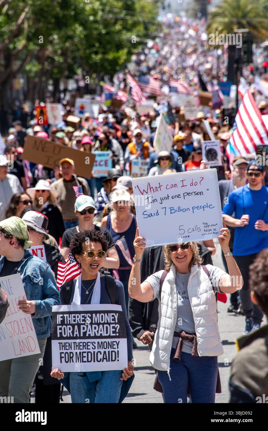 San Francisco, USA. 14th June 2025. Two smiling women are seen in the foreground of thousands of protesters marching down Market Street in San Francisco's massive No Kings Day protest. One holds a sign reading, 'If there's money for a parade? Then there's money for Medicaid!' and the other's reads, ''What can I do? I'm just one person,' said 7 billion people,' followed by a note about 5 calls, the app that helps constituents contact their representatives. Credit: Shelly Rivoli/Alamy Live News - Stock Image