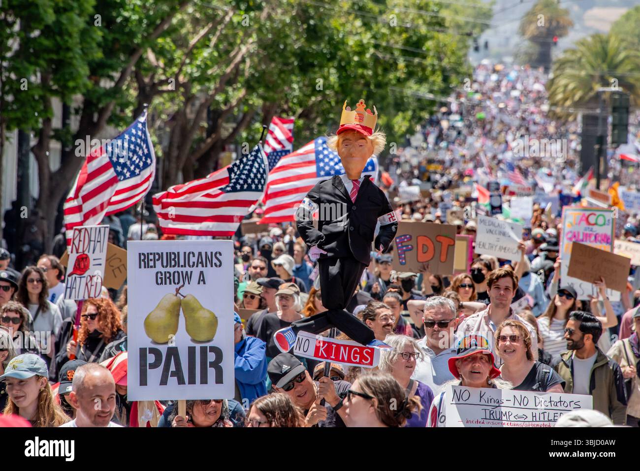 San Francisco, USA. 14th June 2025. Above the massive crowd of protesters marching down Market street, a man carries a humorous effigy of Donald Trump wearing a Burger King crown for the No Kings Day protest in San Francisco. Multiple American flags are seen behind it. Credit: Shelly Rivoli/Alamy Live News - Stock Image