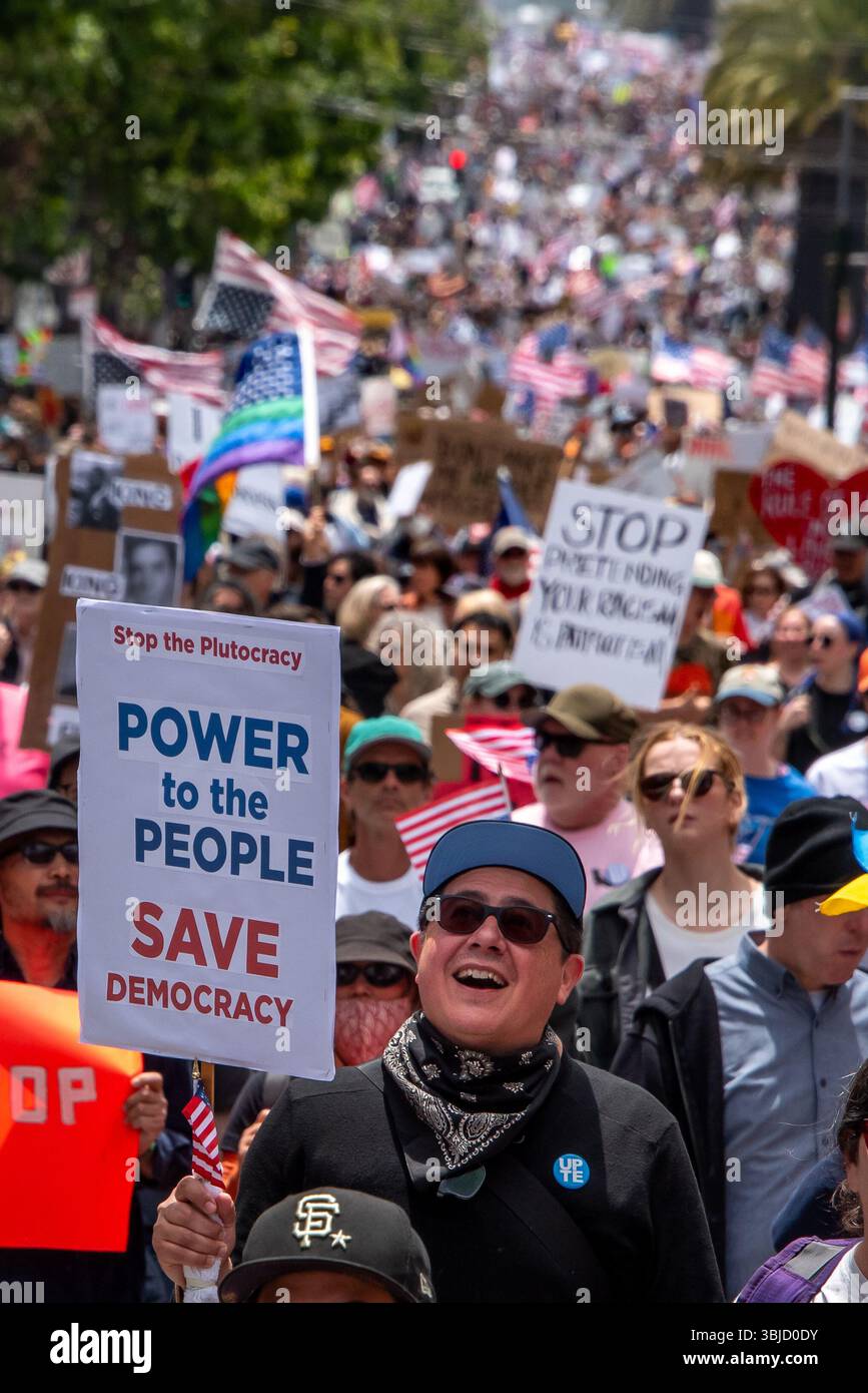San Francisco, USA. 14th June 2025. A protester smiles hopefully while carrying a sign reading, 'Stop the Plutocracy, Power to the People. Save Democracy,' with a crowd of thousands of protesters marching into the distance behind him at the No Kings Day protest in San Francisco. Credit: Shelly Rivoli/Alamy Live News - Stock Image