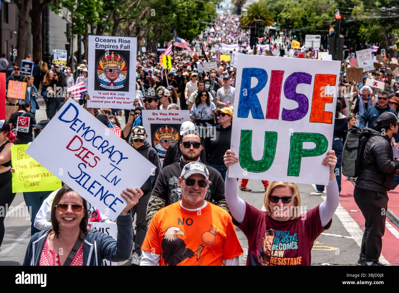 San Francisco, USA. 14th June 2025. Smiling protesters hold up signs reading, 'Democracy dies in silence,' 'RISE UP,' and lines from musicals Hamilton and Les Miserables: 'Oceans rise, Empires fall,' and 'Do you hear the people sing?' Behind them thousands of protesters fill the street into the diestance. Credit: Shelly Rivoli/Alamy Live News - Stock Image
