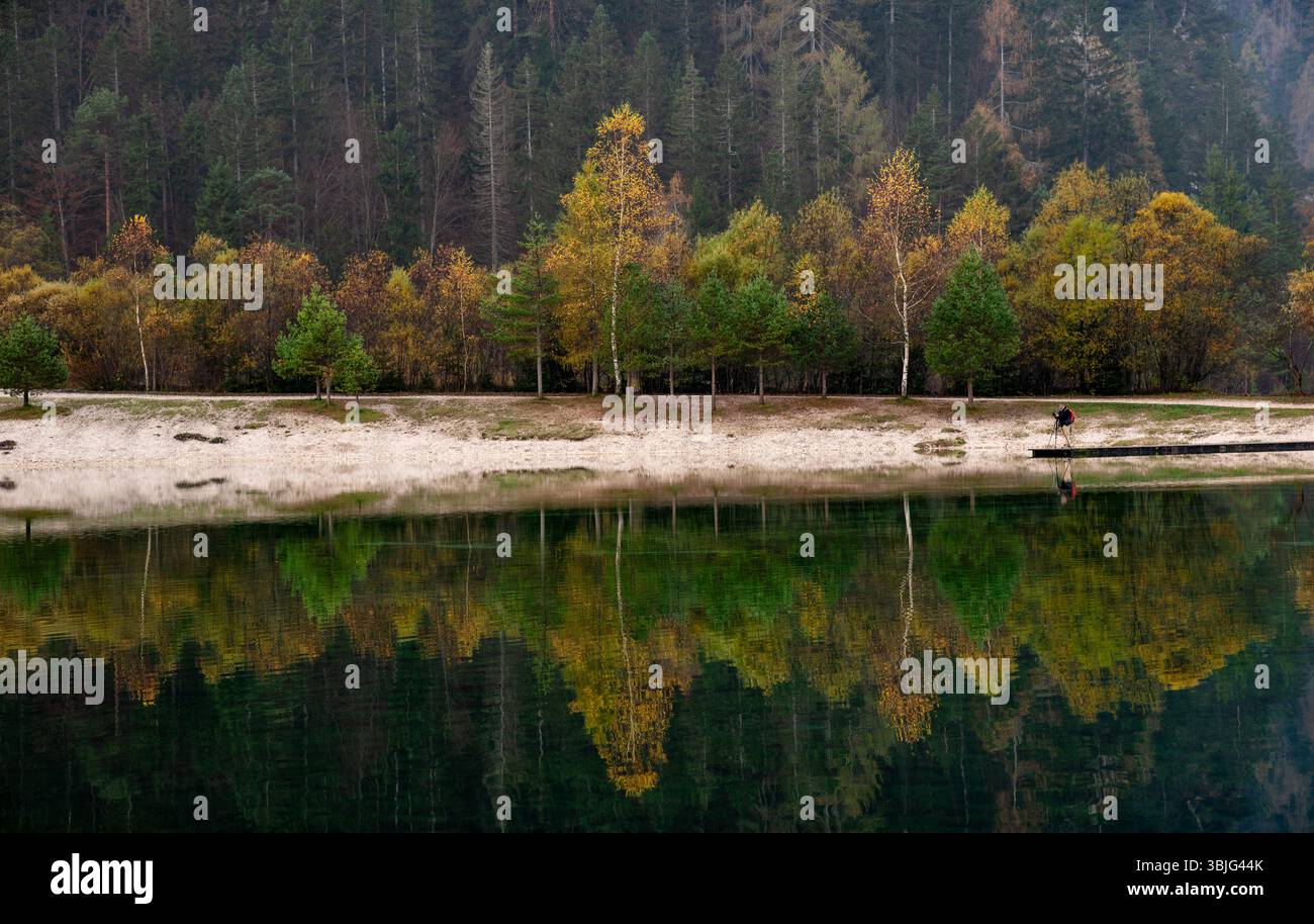 Colorful autumn trees reflecting on the surface of a calm lake with photographer standing on a wooden pier, Lake jasna Slovenia Stock Photo