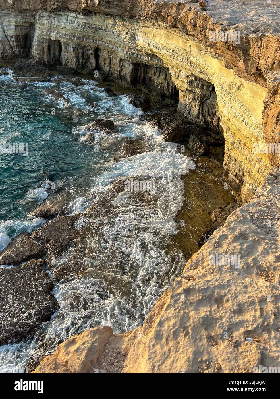 Waves crashing against layered sea cliffs and rock formations at sunset. A dramatic coastal landscape shaped by erosion and time. - Smartphone Captured Stock Image