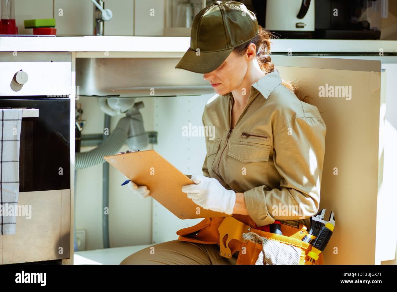 A focused plumber inspects plumbing under a kitchen sink, ensuring everything is working correctly. Stock Photo
