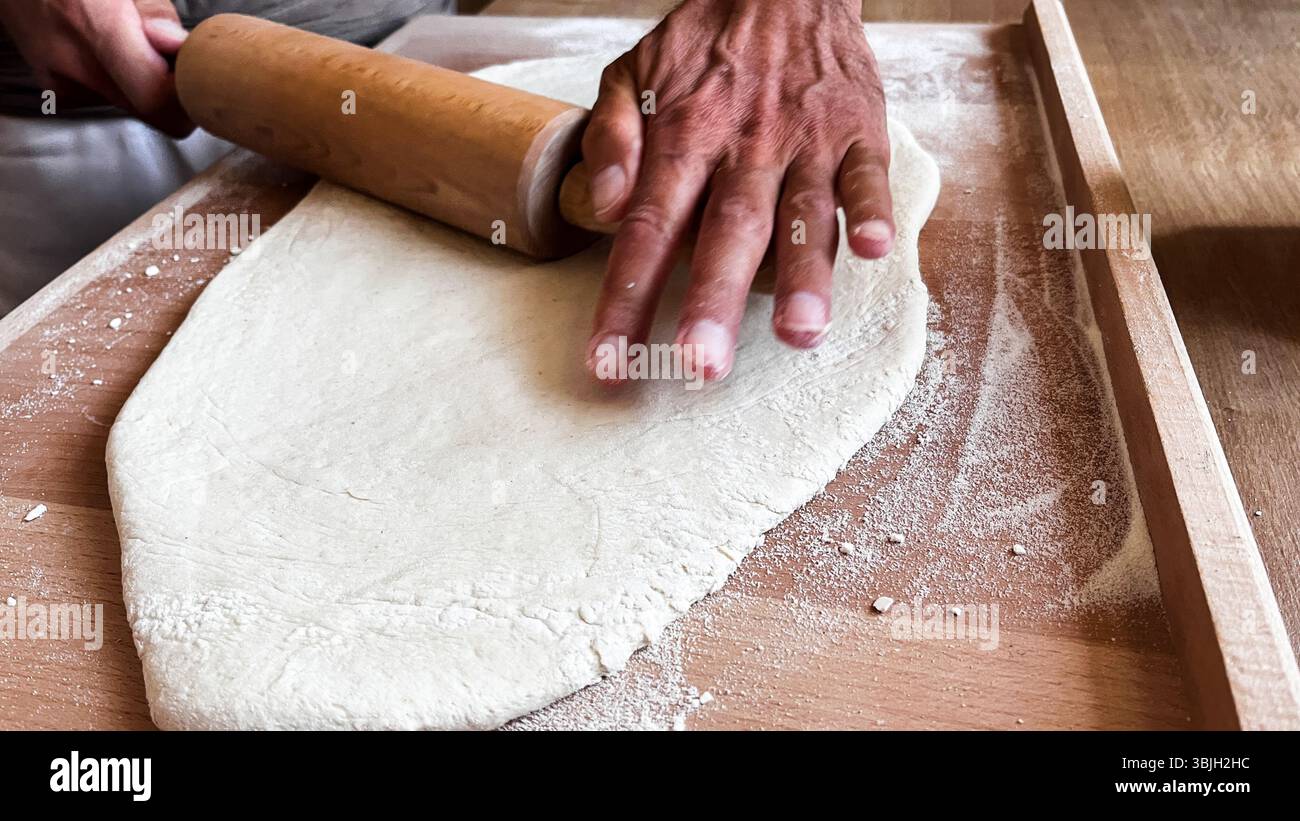 Close up of man's hands rolling dough on wooden table with wooden rolling pin. Homemade baking, step by step cooking class, preparation, pastry Stock Photo