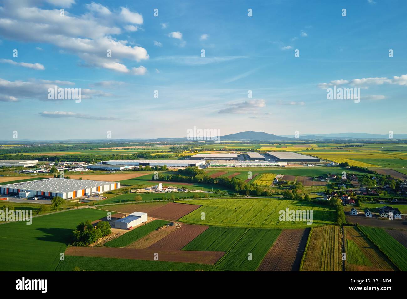 Aerial view of industrial warehouse complex near green agricultural fields and small village. Modern logistics infrastructure in rural landscape Stock Photo