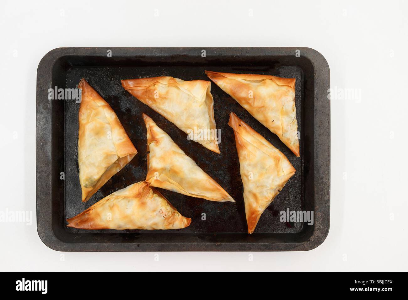 Golden brown, triangular pastries on a baking tray arranged in a pattern against a plain white background Stock Photo