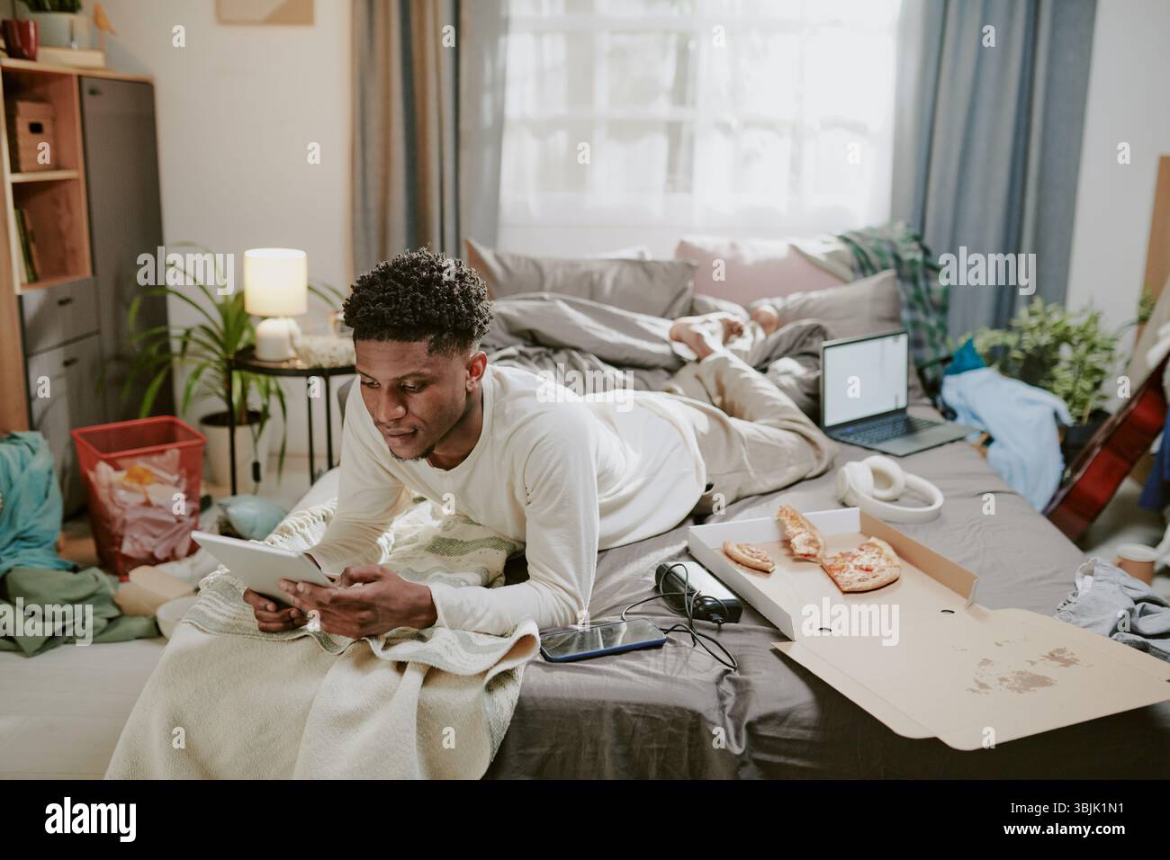 Young Man Lying on Bed Using Tablet in Cluttered Room Stock Photo