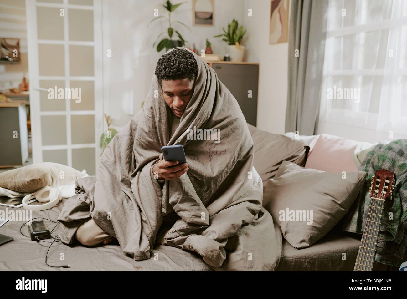 Man Wrapped in Blanket Using Smartphone Indoors Stock Photo