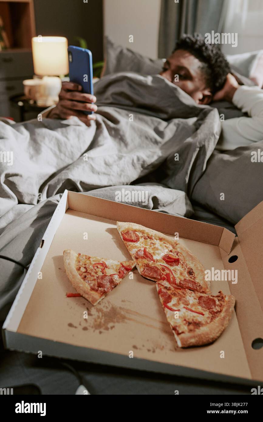 Relaxing in Bed Enjoying Pizza While Using Mobile Device Stock Photo