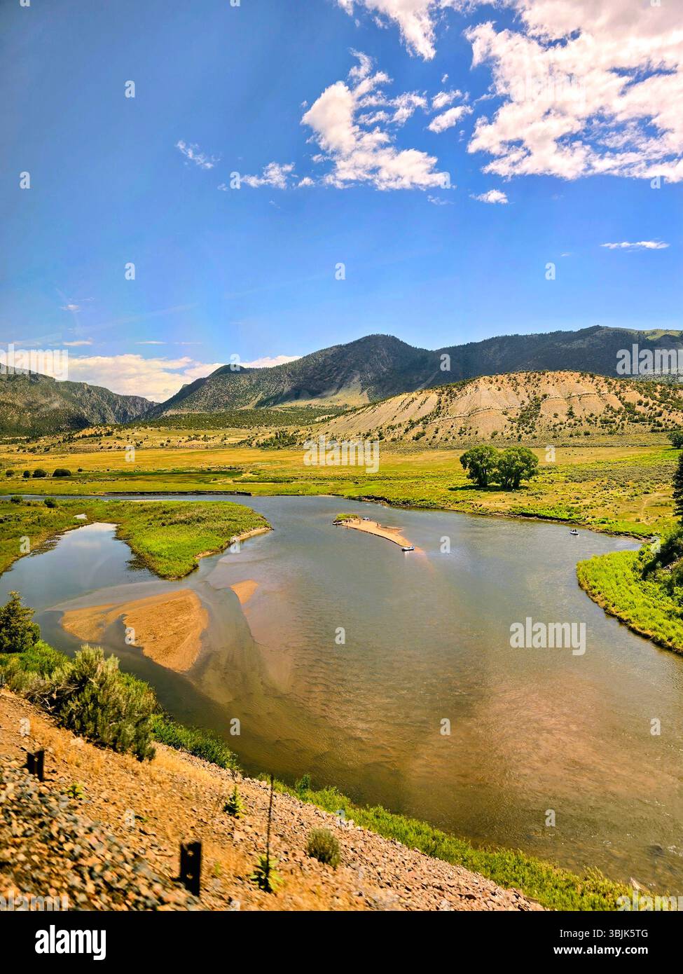 Winding river bend below rugged mountain slopes and forested cliffs in the Colorado backcountry, under bright summer skies. - Smartphone Captured Stock Image