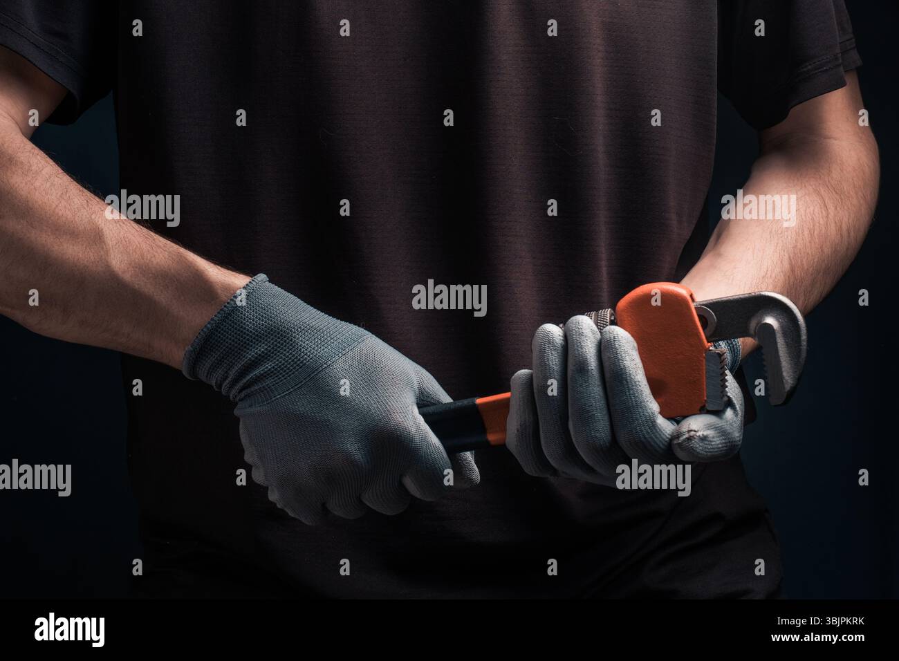 Plumbers hands in protective gloves holding pipe wrench on dark background Stock Photo
