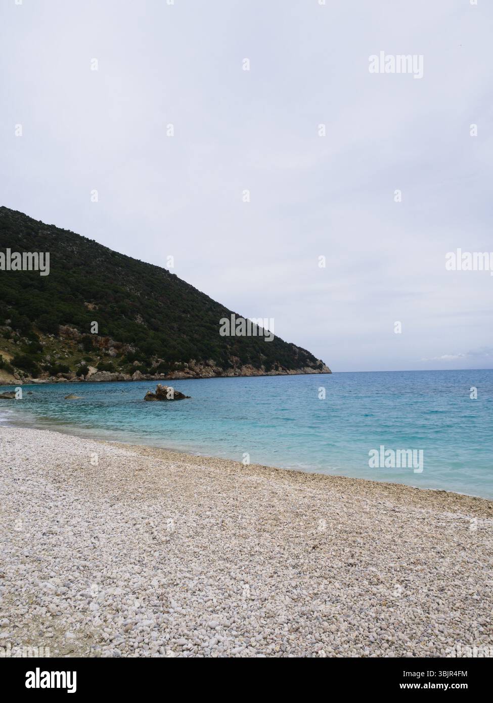 A minimalist gravel beach in Kefalonia under a cloudy sky with a calm sea and visible coastline, ideal for layout or design. Stock Photo